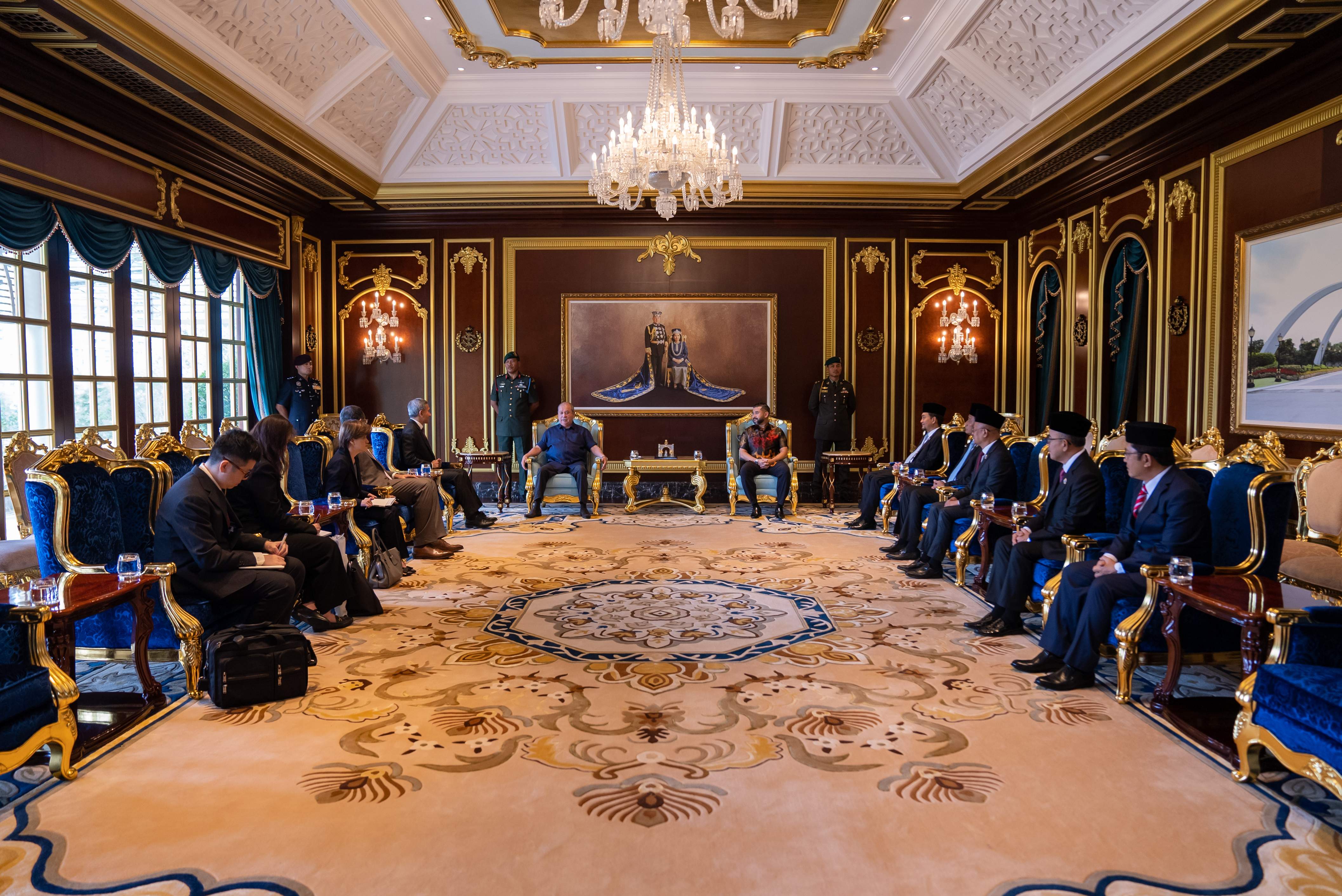 Ornate room with meeting. Men in suits sit in blue/gold chairs facing forward.
